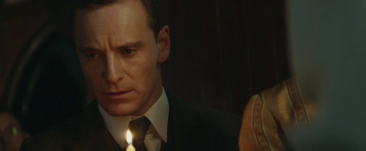 The Light Between Oceans (2016) movie still: closeup — The interior of a dimly lit, dark wood church at night. A man in a suit looks down at the…
