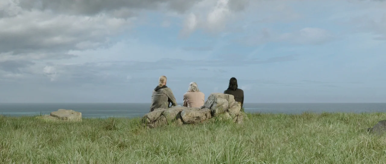 Thor: Ragnarok (2017) movie still: wide — A grassy coastal ridge overlooks a calm ocean under a cloudy sky. Three people sit together on a cluster…
