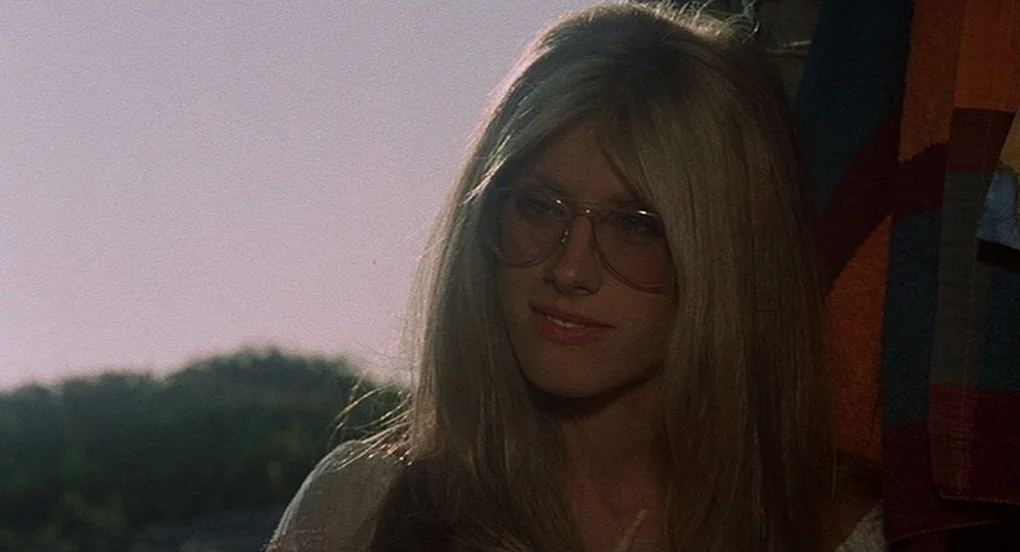 Vanishing Point (1971) movie still: closeup — A woman stands outdoors in natural daylight with a soft-focus background. She wears large glasses…