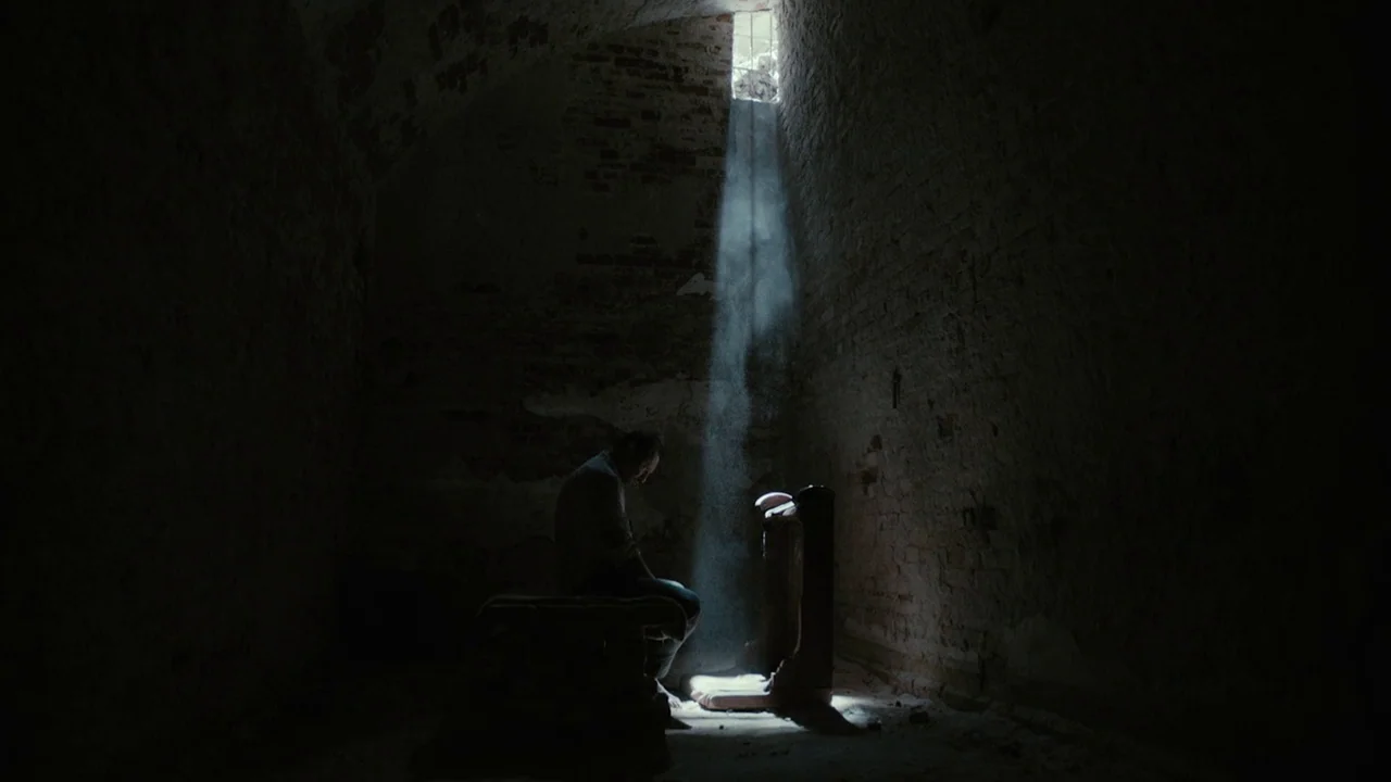 The Field Guide to Evil (2018) movie still: wide — A dark stone cell features rough brick walls and a single high, barred window. A man sits alone…