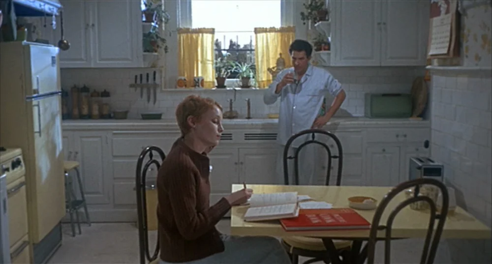 Rosemary’s Baby (1968) movie still: medium — A vintage 1960s residential space features yellow appliances and white wood cabinets. A woman sits at a…