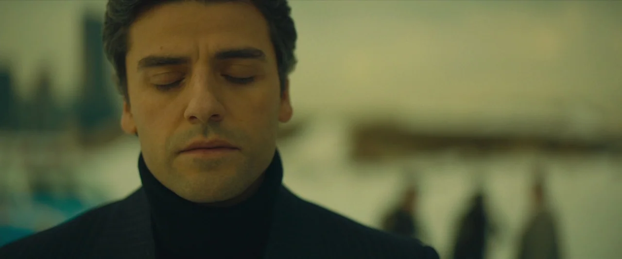 A Most Violent Year (2014) movie still: closeup — An outdoor waterfront setting with blurred figures in the background. A man stands with his eyes…