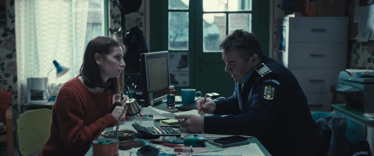 R.M.N. (2022) movie still: medium — A cluttered police office with floral wallpaper, stacks of paper, and a computer monitor. A woman sits across…