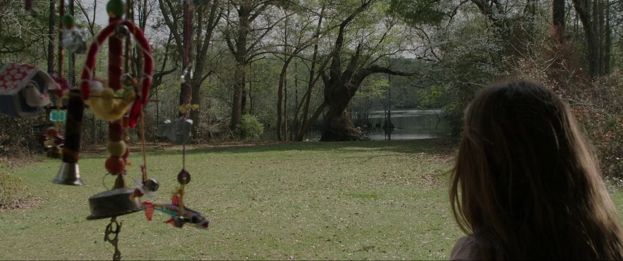 The Conjuring (2013) movie still: medium — A grassy clearing sits between a residential yard and a calm, dark lake. A hanging mobile of assorted…
