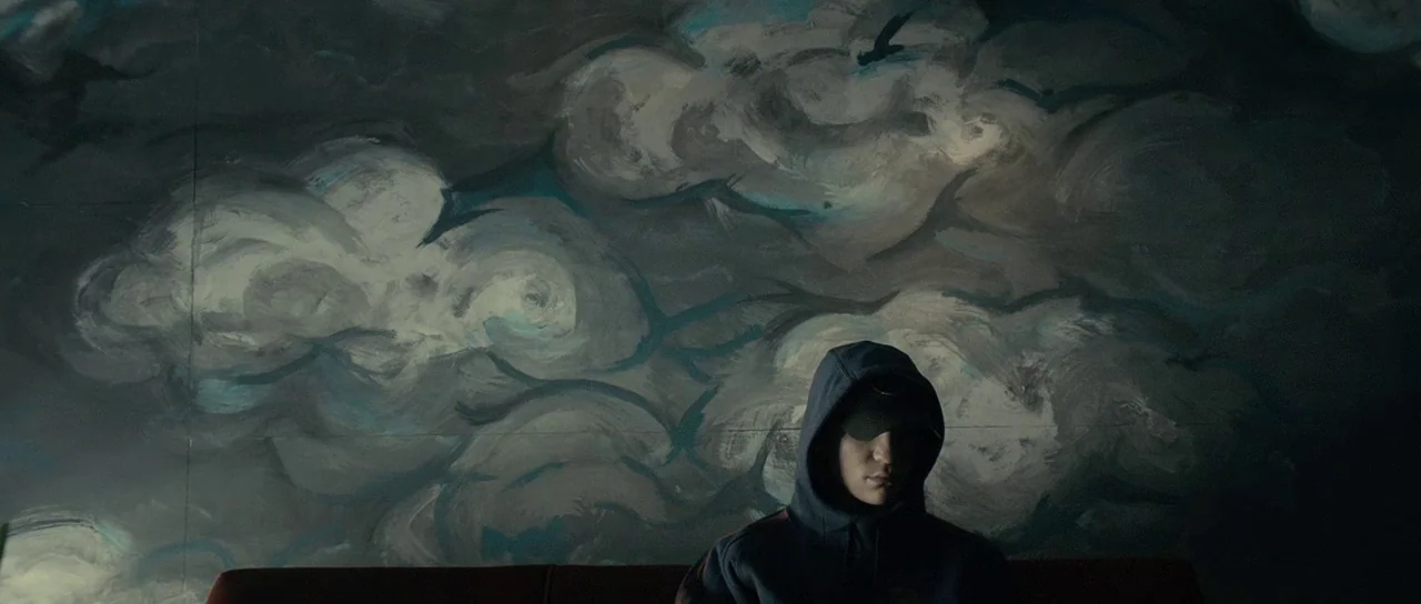 The Imposter (2012) movie still: medium — A dimly lit room features a wall decorated with a large mural of clouds. A man wearing a hooded sweatshirt…