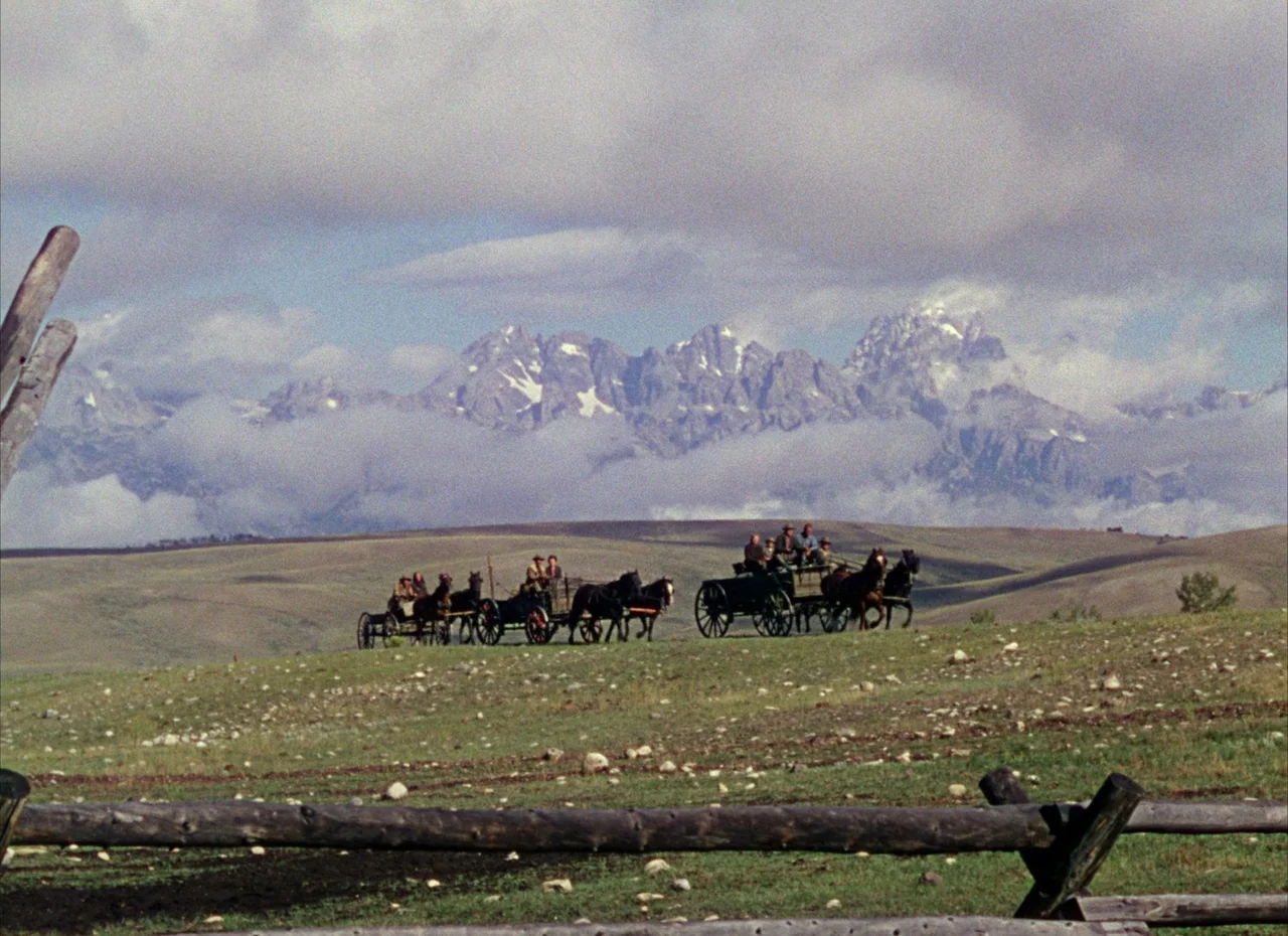 Shane (1953) movie still: wide — A vast, grassy prairie sits beneath jagged mountains partially obscured by clouds. Two horse-drawn carriages move…