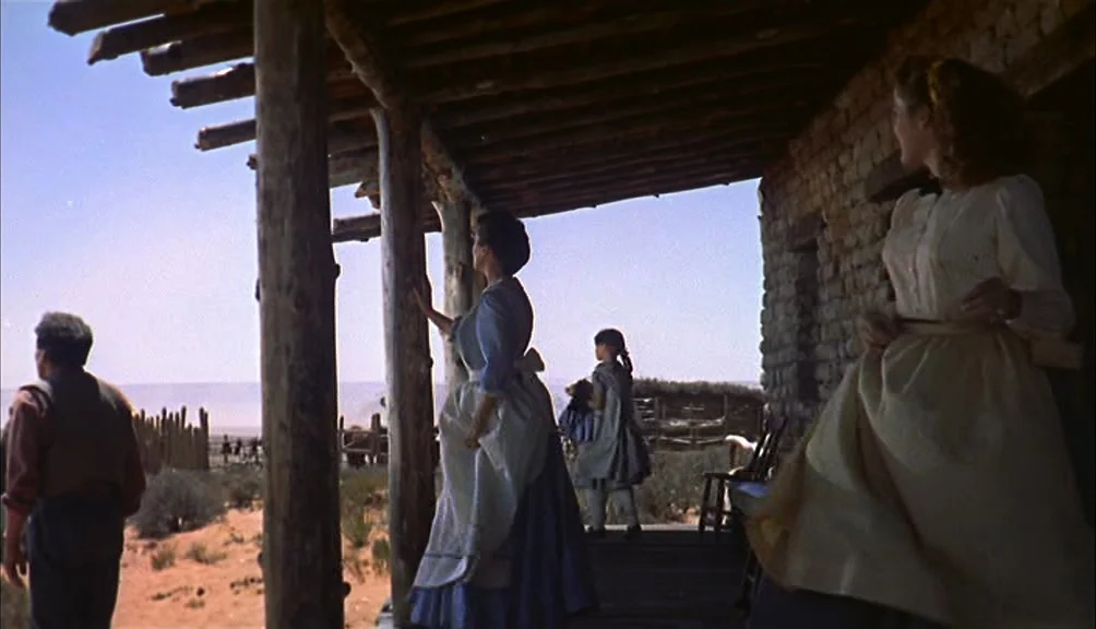 The Searchers movie still: wide — A rustic adobe dwelling with a wooden porch overlooks a barren desert landscape. A family group stands on the…