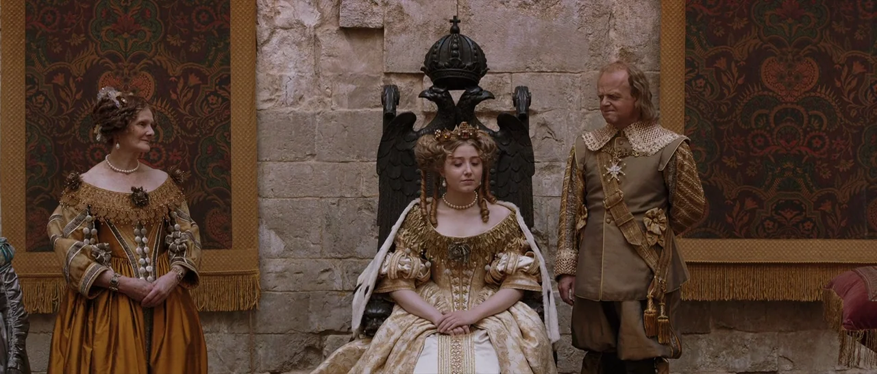 Tale of Tales (2015) movie still: fullbody — A stone-walled interior features ornate tapestries hung on either side of a large throne. A young queen…