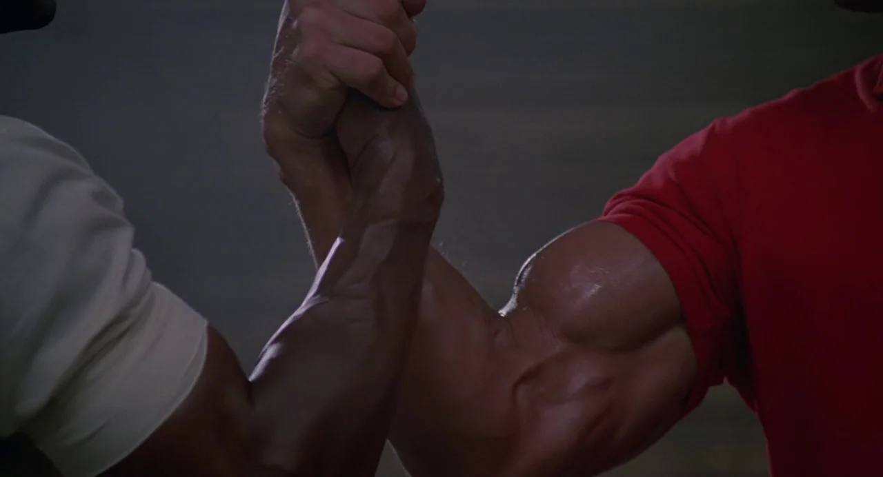 Predator (1987) movie still: closeup — Two muscular arms grip each other tightly in a firm handshake gesture. One arm belongs to a person wearing a…