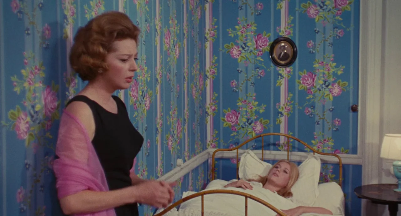 The Umbrellas Of Cherbourg (1964) movie still: medium — A bedroom decorated with blue floral wallpaper and a gold-colored metal bed frame. One woman…