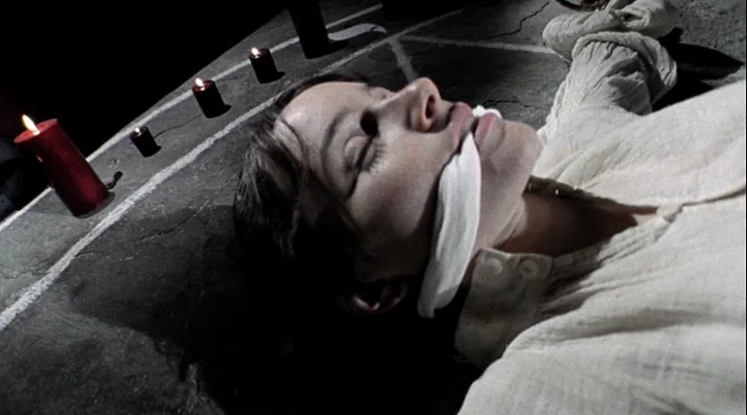 The House Of The Devil (2009) movie still: closeup — A dark, uneven stone surface is marked with white chalk lines and glowing candles. A young…