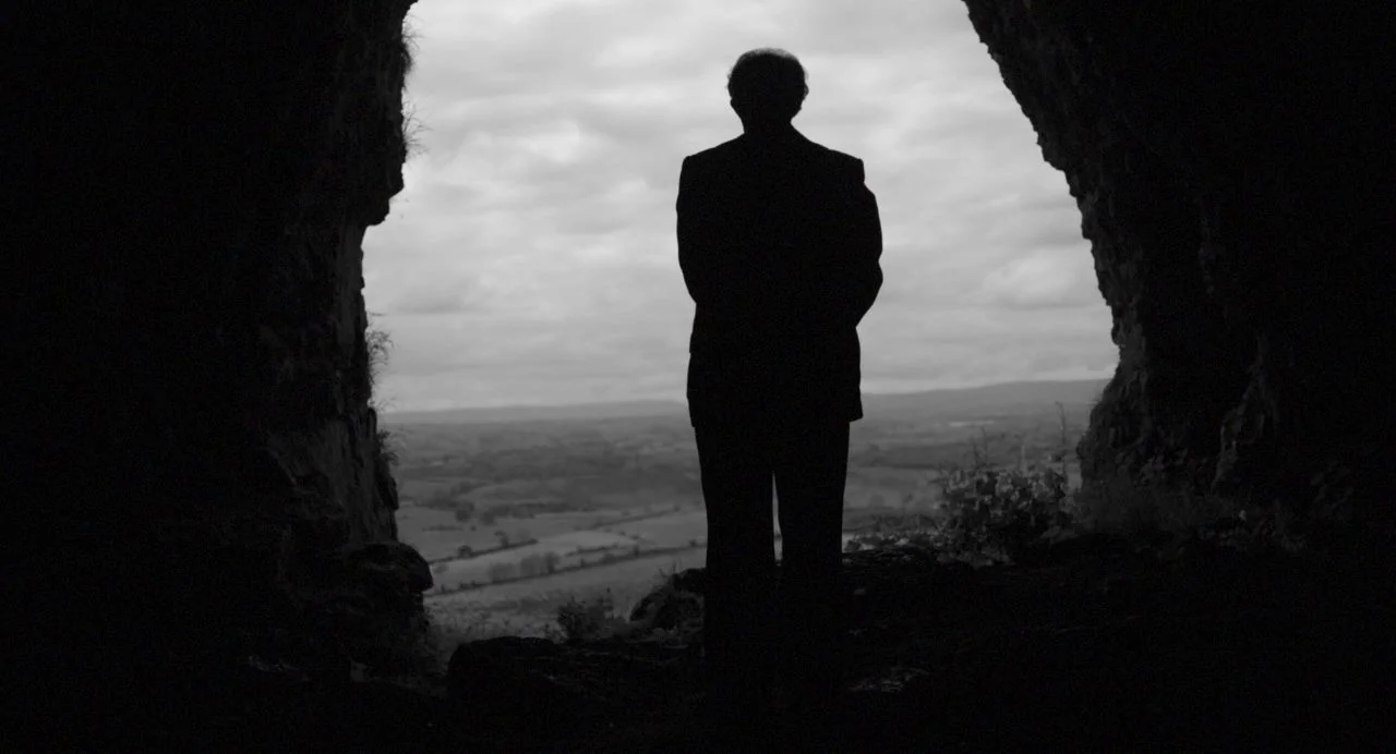 Song of Granite (2017) movie still: fullbody — A man stands inside a rocky cave entrance looking out over a rural landscape. The bright overcast sky…