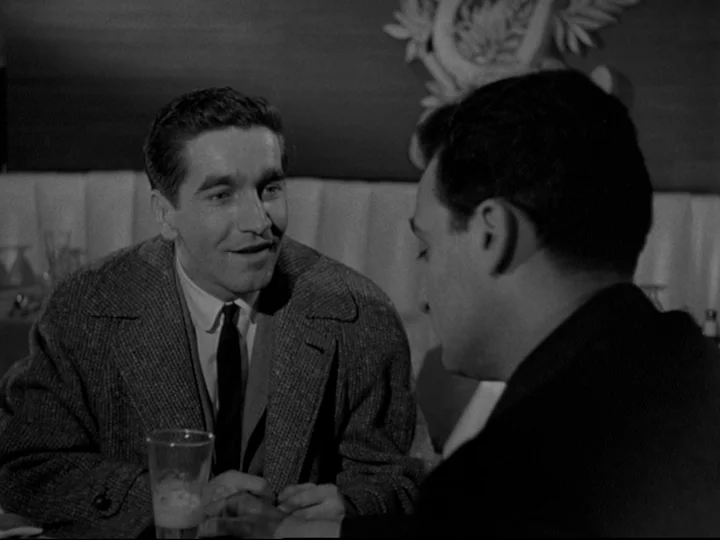 Blast Of Silence (1961) movie still: medium — A dimly lit diner booth features a padded wall behind two men. The men sit opposite one another at a…