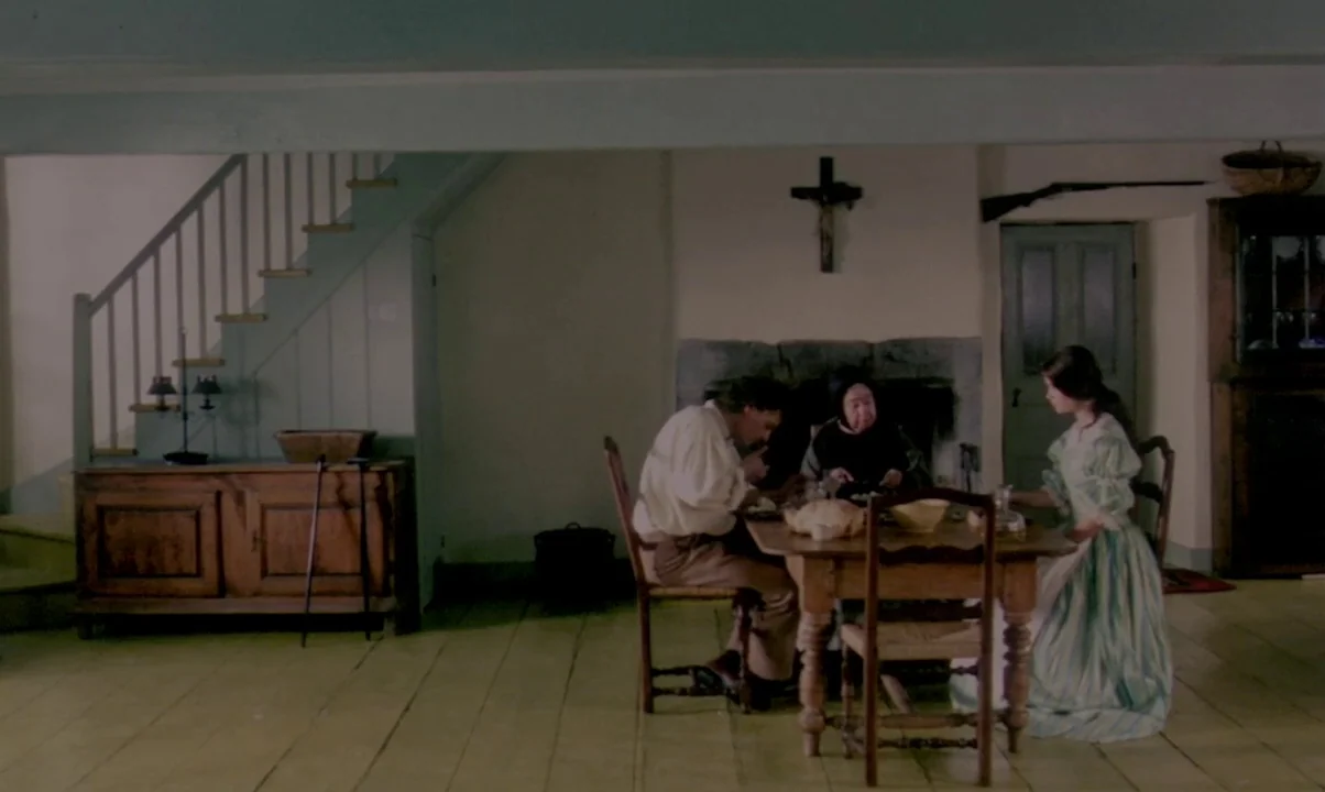 Kamouraska (1973) movie still: wide — A rustic 19th-century dining room features wooden plank flooring and a stone fireplace. Three people sit at a…