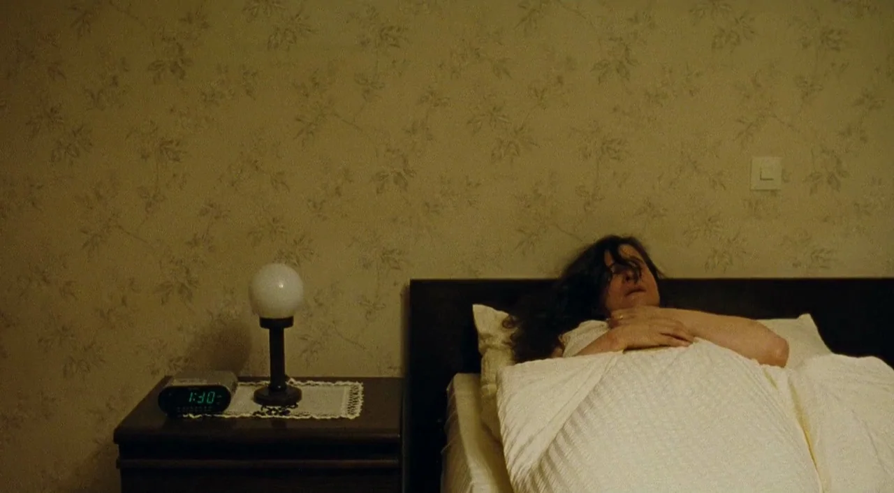 Paradise: Faith (2012) movie still: medium — A woman lies under white blankets in a room with faded floral patterned wallpaper. She rests with her…