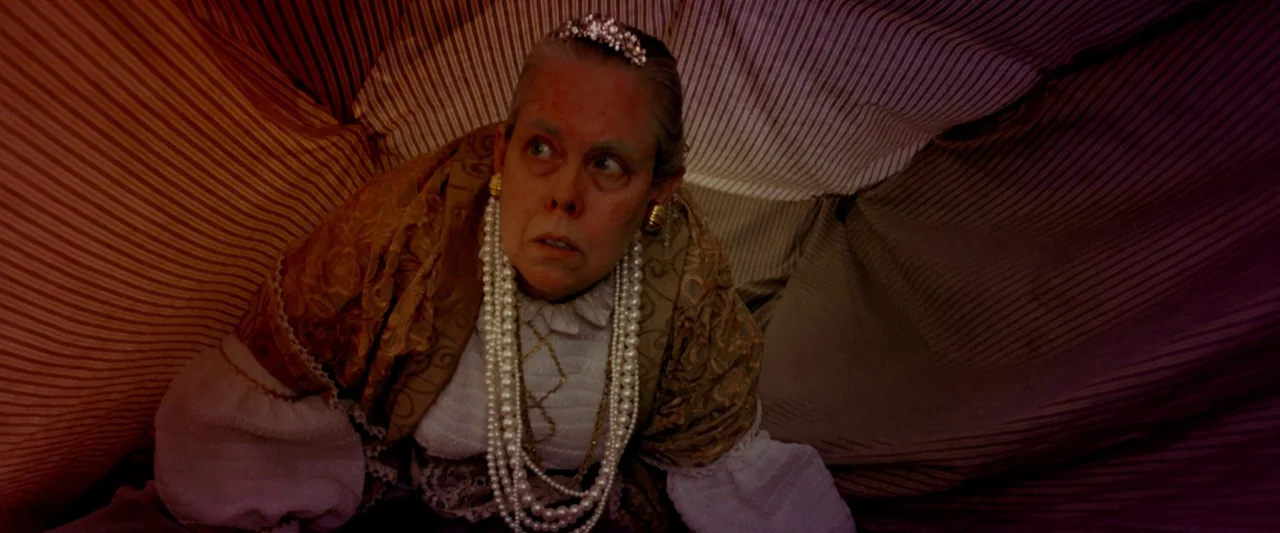 Moon Garden (2022) movie still: medium — A woman sits beneath a draped striped tent-like structure of brown and red fabric. She looks upward while…