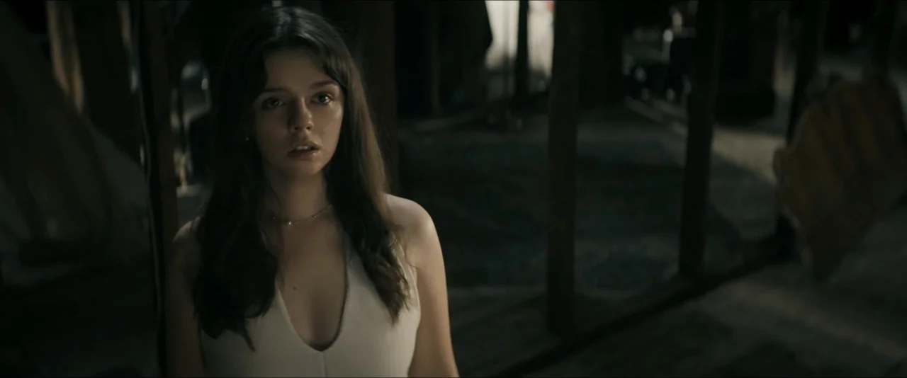 It Feeds (2025) movie still: medium — A dimly lit room with exposed wooden beams and structural supports visible in darkness. A young woman with…