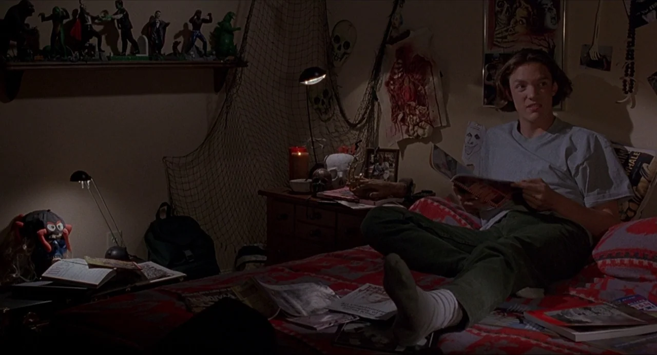 Serial Mom (1994) movie still: medium — A dimly lit room filled with horror-themed memorabilia including monster figurines and anatomical posters. A…