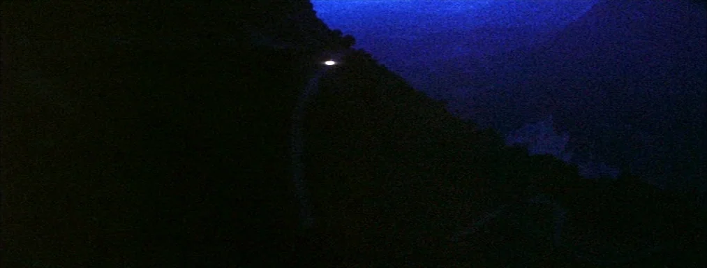 Japón (2002) movie still: establishing — A dark, mountainous landscape is rendered in deep blue tones at night. A single bright headlight traverses…