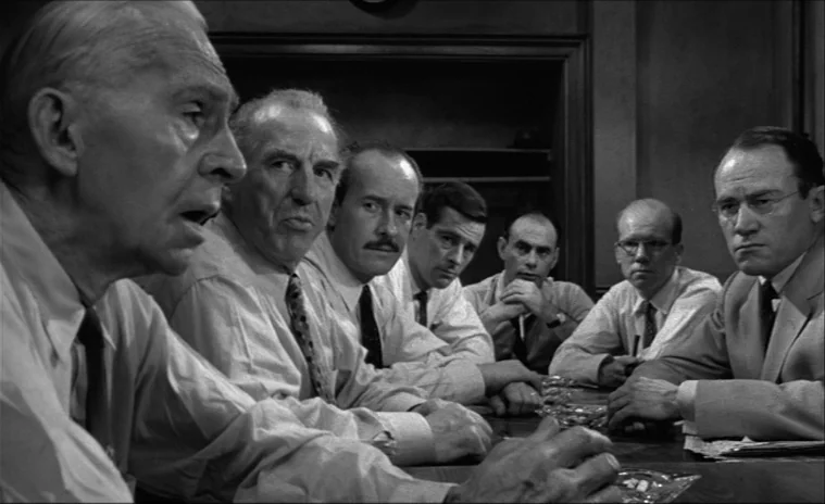 12 Angry Men (1957) movie still: medium — A group of seven men sit around a wooden table in a dimly lit office. They are engaged in a tense…