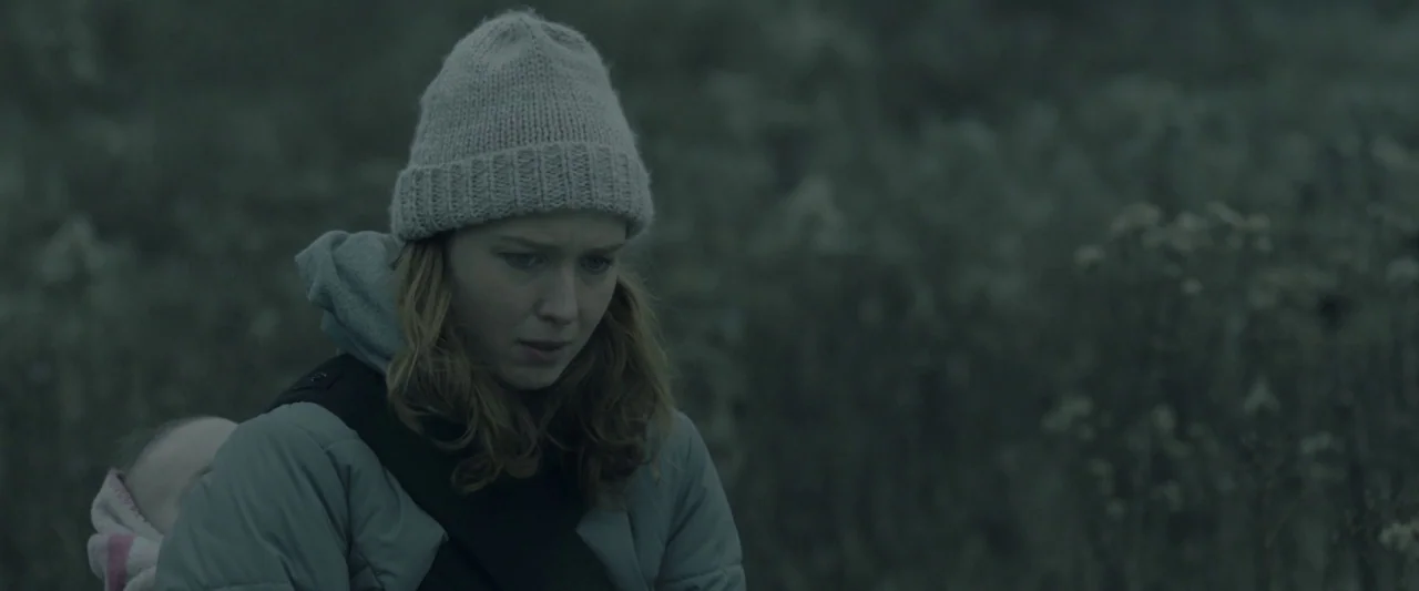 Here Alone movie still: medium — A woman stands in a desolate field with tall, dried-out vegetation under an overcast sky. She holds an infant in a…