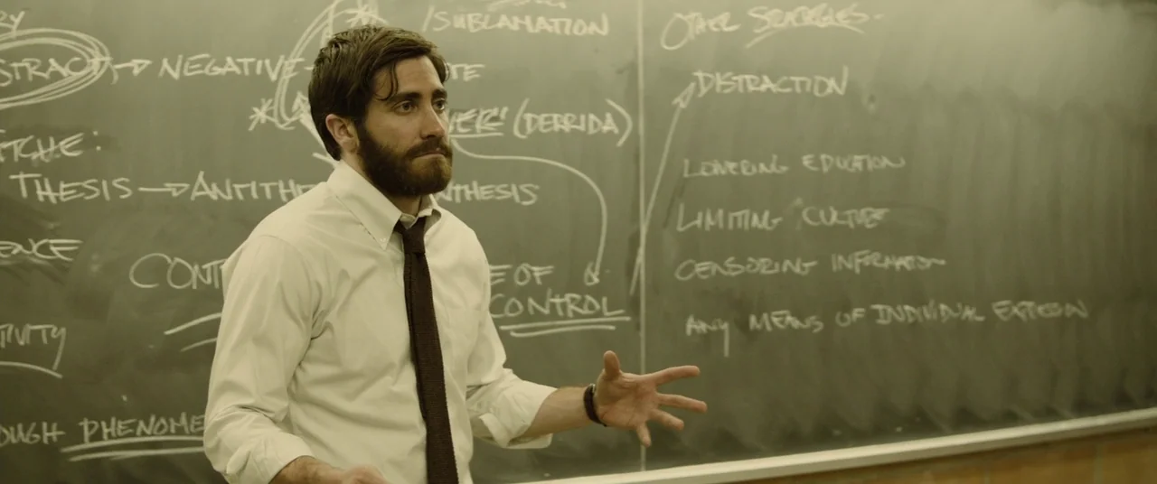 Enemy (2013) movie still: medium — A man stands in front of a large chalkboard covered in handwritten academic notes. He gestures with his hand…