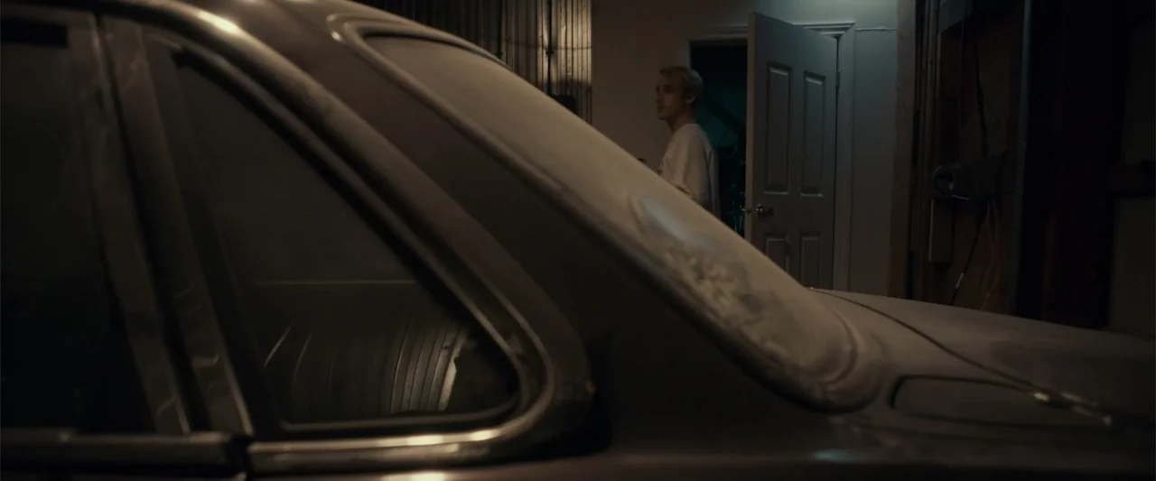 Jump, Darling (2022) movie still: medium — A dimly lit garage interior features a dusty car in the immediate foreground. A young man stands near an…
