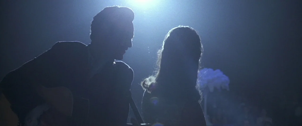 Walk The Line (2005) movie still: medium — A dark concert stage with a strong backlight obscuring the subjects. A man plays an acoustic guitar while…