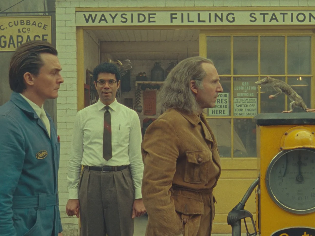 The Rat Catcher (2023) movie still: medium — A vintage 1950s filling station with a yellow facade and clear signage. Three men stand together in…