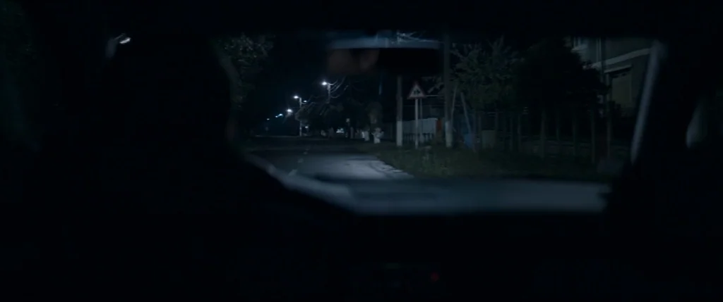 Graduation (2016) movie still: wide — A dark, empty road is seen through the windshield of a moving vehicle. Dim streetlights provide sparse…