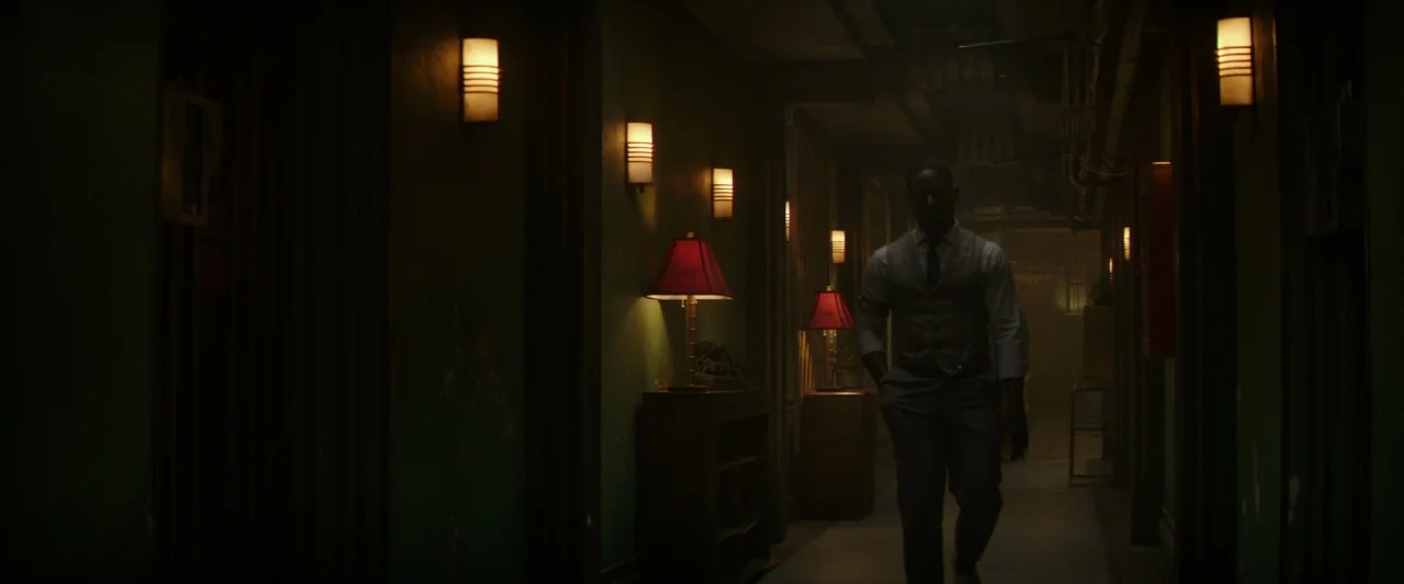 Hotel Artemis (2018) movie still: fullbody — An interior building hallway with dark green walls is lit by multiple small sconces. A man in a formal…