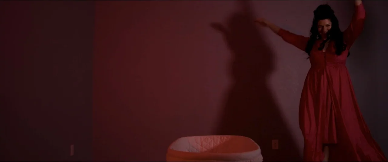 Amityville Cult (2021) movie still: fullbody — A sparsely furnished room with a bassinet positioned in the foreground center. A woman in a red gown…