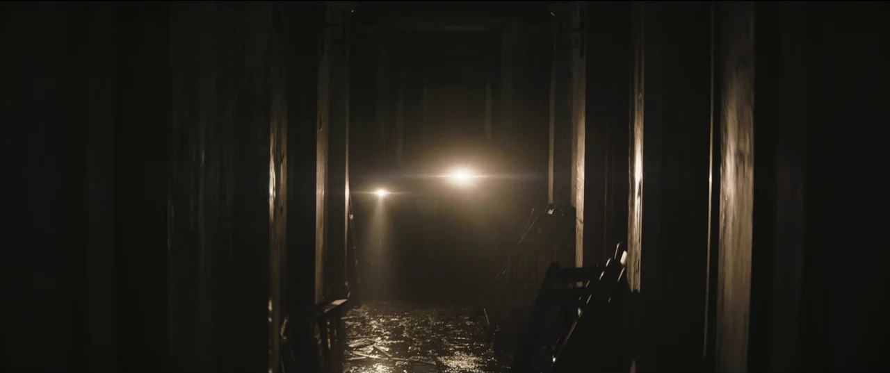 The Batman (2022) movie still: wide — A dark, narrow corridor features debris scattered across the wet floor surface. Two intense beams of light cut…