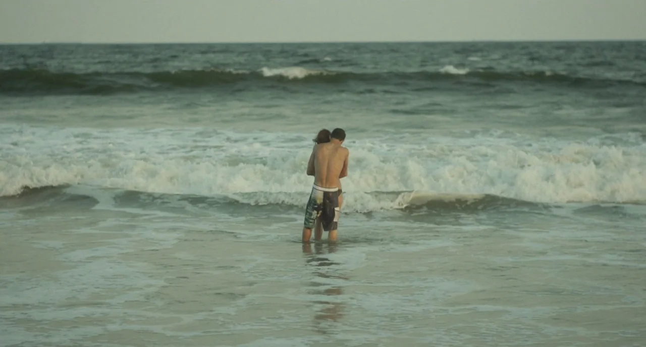 It Felt Like Love (2013) movie still: wide — Two individuals stand in shallow ocean water while waves break around their legs. They are facing away…