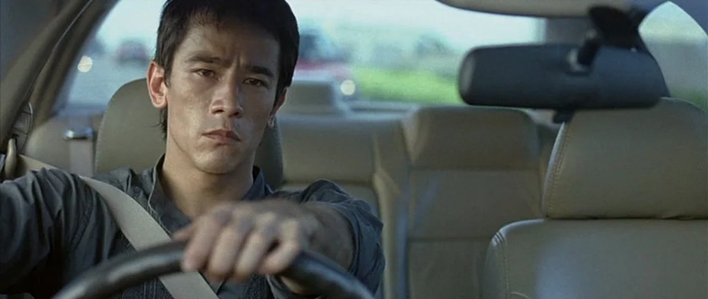 Boarding Gate (2007) movie still: medium — The interior of a modern car with light-colored upholstery and headrests. A young man with dark hair is…