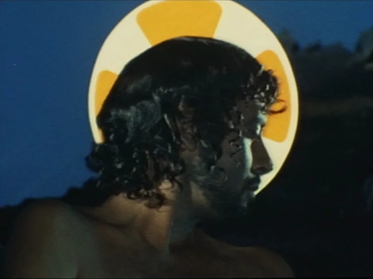 Salome (1972) movie still: closeup — A person is in water at night against a deep blue background. A large yellow graphic of a life buoy appears…