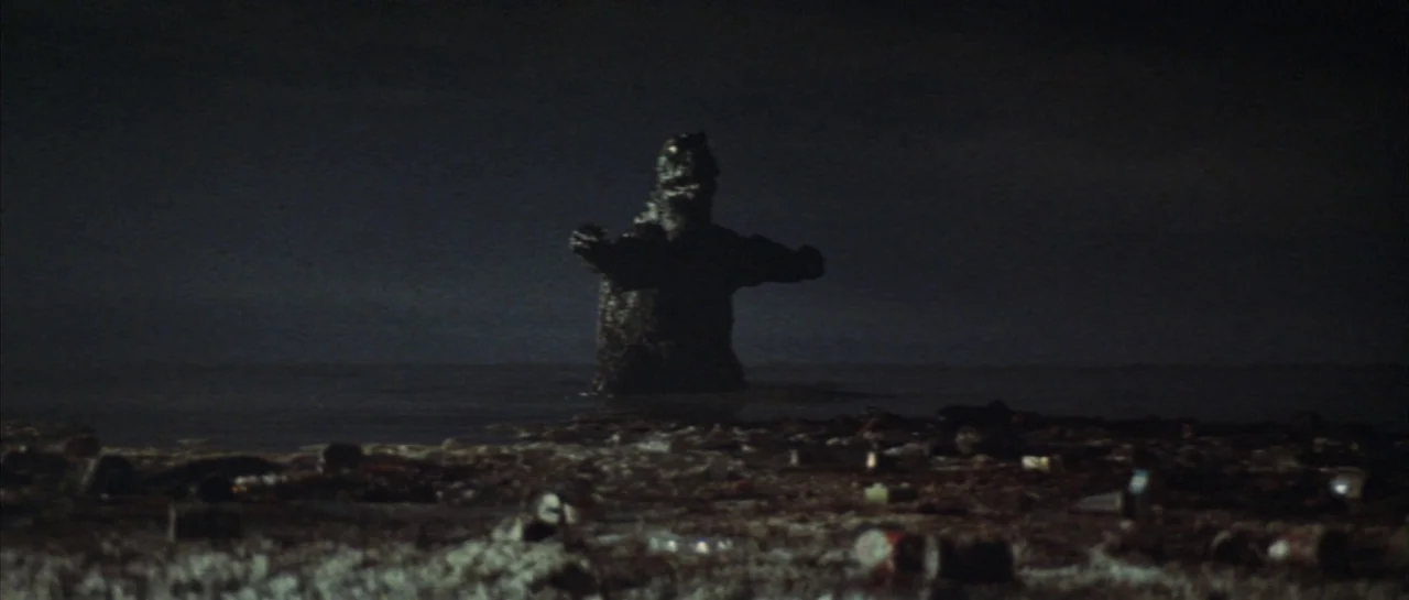 Godzilla Vs Hedorah (1971) movie still: wide — A dark night at a polluted beach littered with debris and scattered trash. The large reptilian…