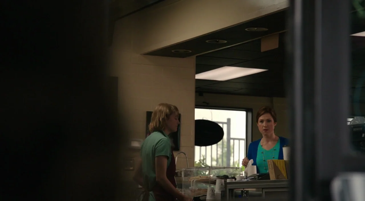 Laggies (2014) movie still: medium — A mundane commercial service counter with neutral wall paint and a large glass window. A woman with short…
