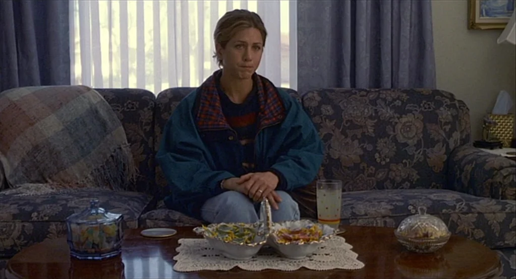The Good Girl (2002) movie still: medium — A residential living room features a patterned sofa with a plaid throw blanket. A woman sits on the sofa…