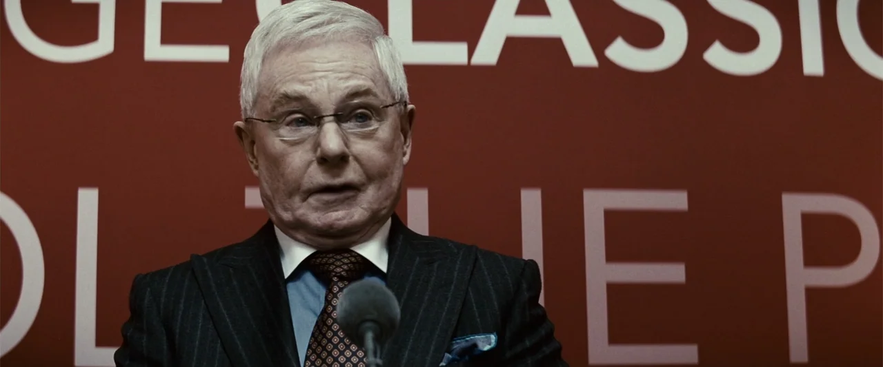 Hereafter (2010) movie still: medium — An elderly man stands in front of a deep red wall with white lettering. He speaks into a microphone at a…