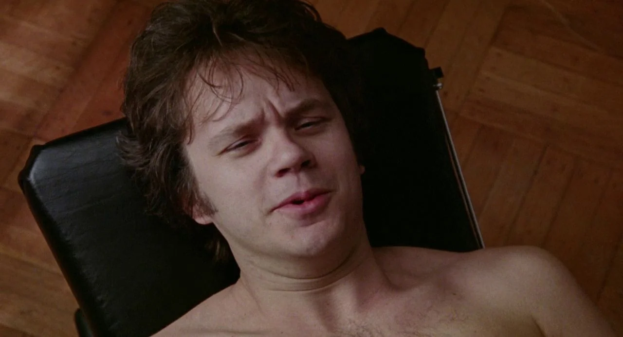 Jacob’s Ladder (1990) movie still: closeup — A man lies supine on a black-upholstered chair above a light-toned wooden floor. He stares upward with…