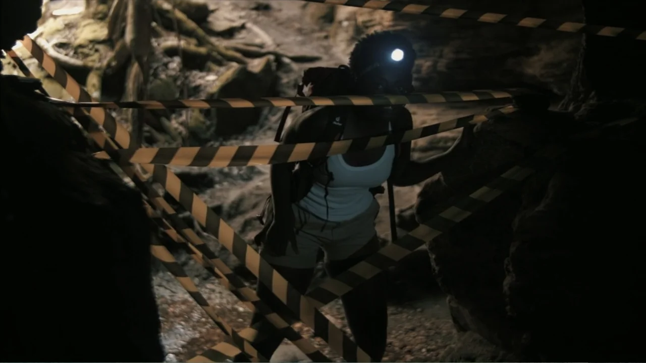 The Grotto (2022) movie still: medium — A rocky, dimly lit cave interior features uneven ground and textured stone walls. A woman wears a headlamp…