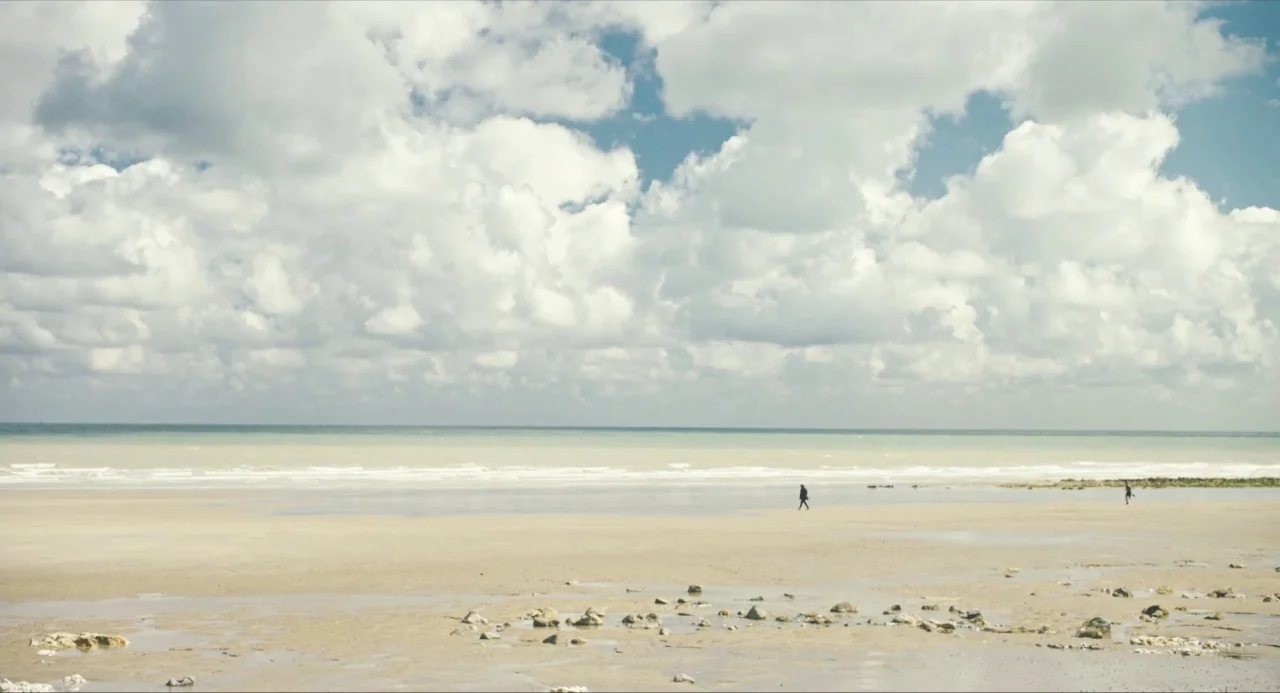 Submergence movie still: wide — A vast, flat sandy beach extends to a distant horizon line under a cloudy sky. Two small, isolated human figures…