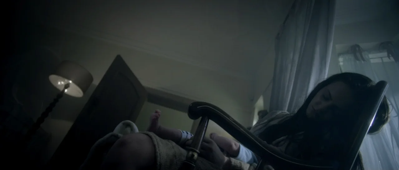 The Lullaby (2019) movie still: medium — A dimly lit nursery features a wooden rocking chair placed near a draped window. A woman sits in the chair…