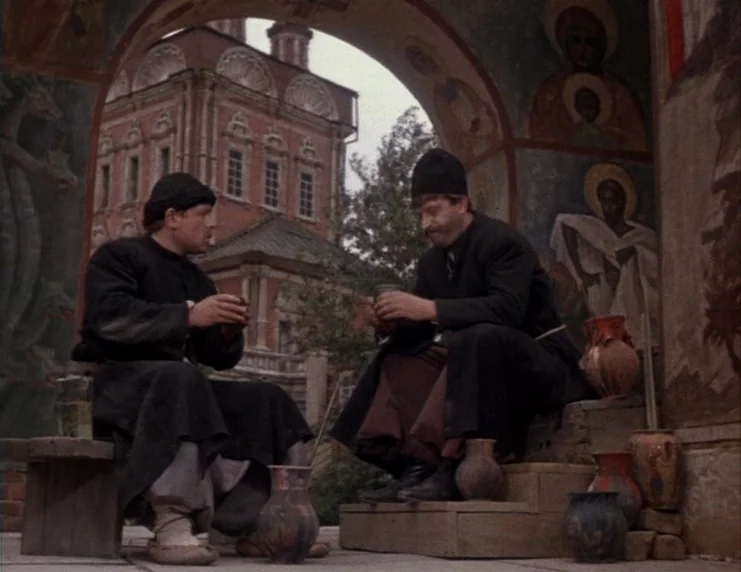 Viy (1967) movie still: fullbody — Two men in dark monastic robes sit beneath a stone archway adorned with colorful religious iconography. They hold…
