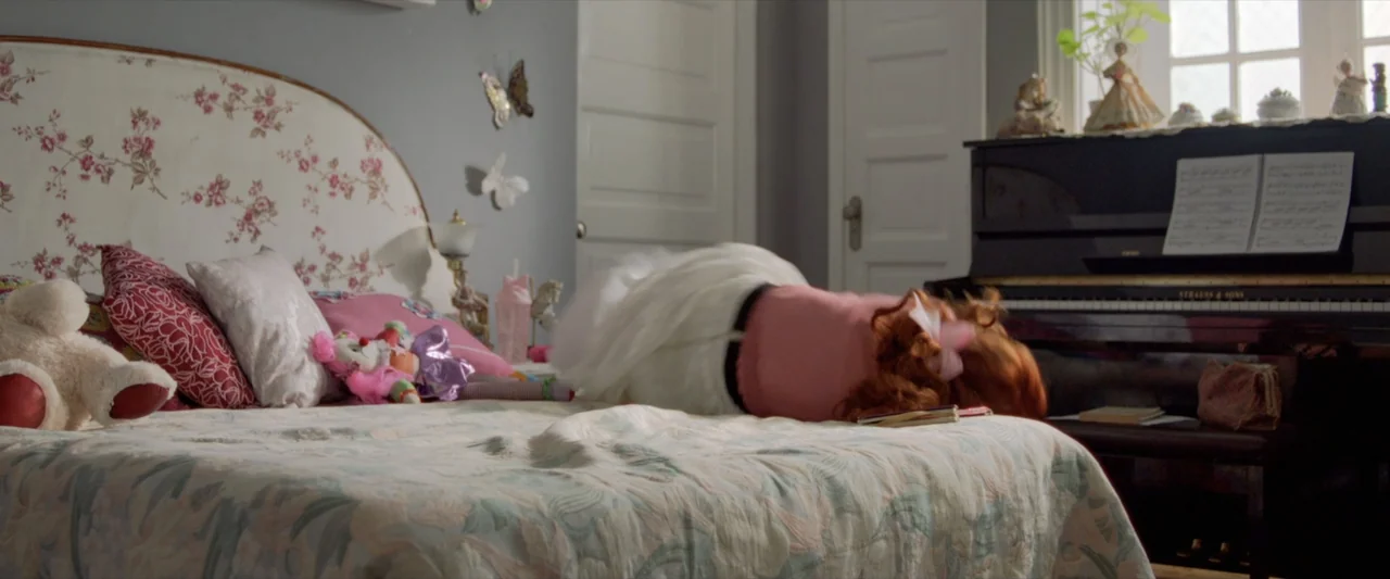 Like or Die (2023) movie still: medium — A girl's bedroom features a patterned bedspread, floral headboard, and an upright piano. A young girl with…