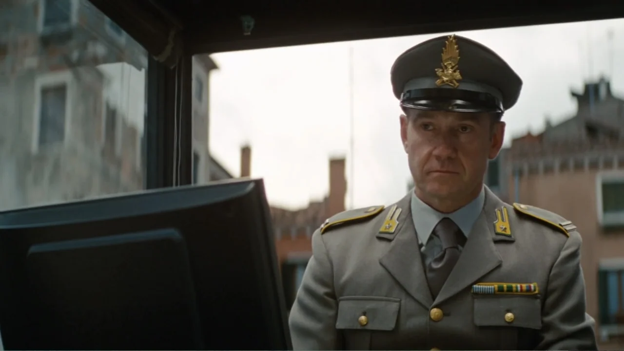 The Tourist (2010) movie still: medium — A man in a military uniform sits inside a small booth with a large window. He stares forward at a computer…