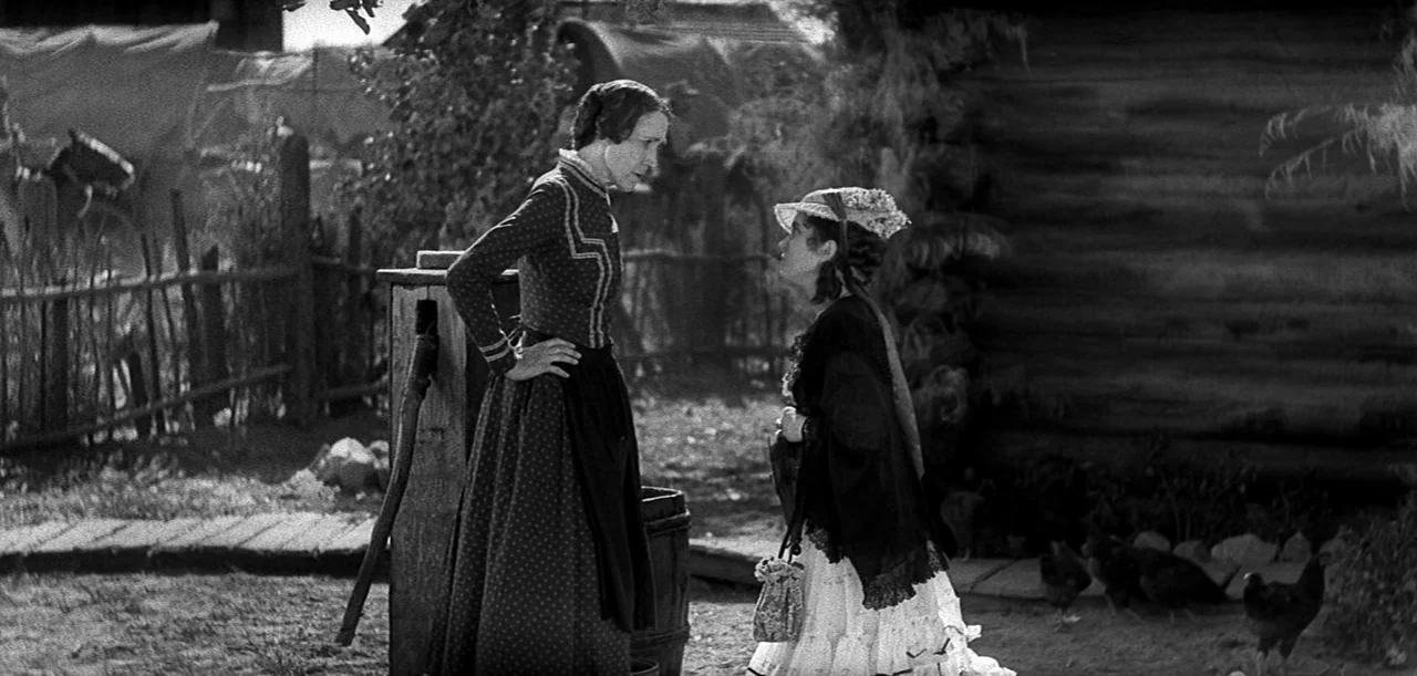 The Big Trail (1930) movie still: fullbody — A rural farmyard exterior features a weathered log structure and a wooden fence. Two women stand facing…