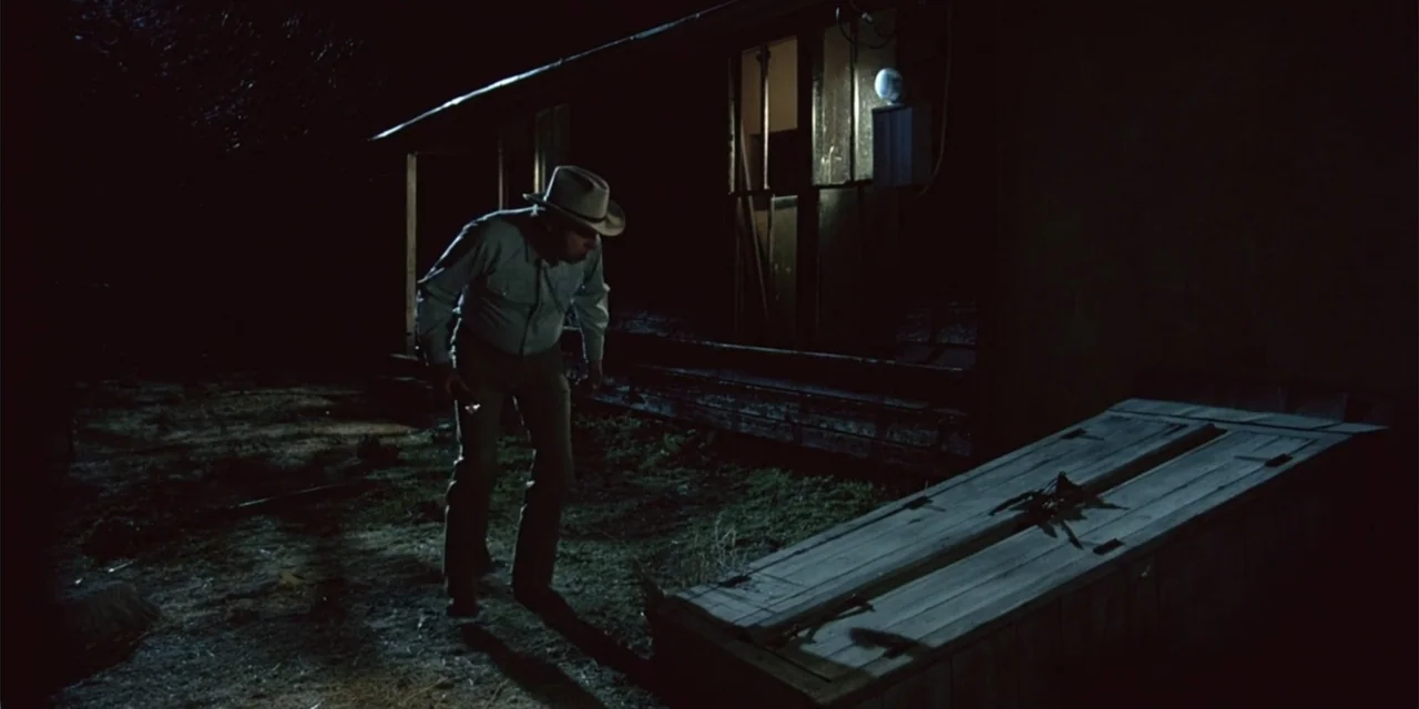 El sótano (2024) movie still: fullbody — An exterior nighttime view of a rural building with a dark, grassy yard. A man in a hat stands near a…