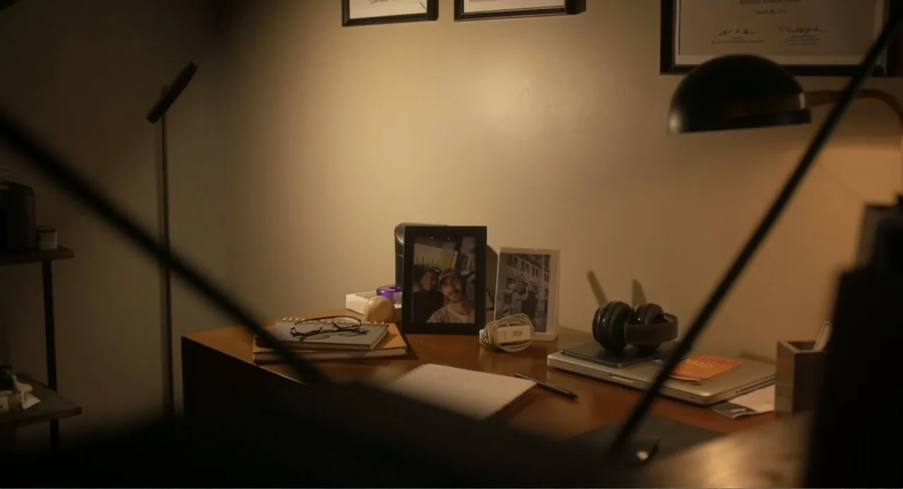 Straight (2024) movie still: medium — A wooden office desk sits under a desk lamp in a dimly lit room. Various personal items and documents are…